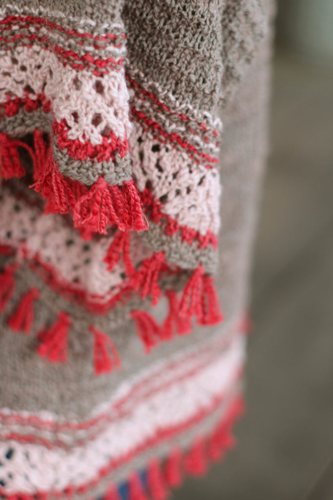 Close up of knitted sleeve edge showing pink lace border and short red fringe