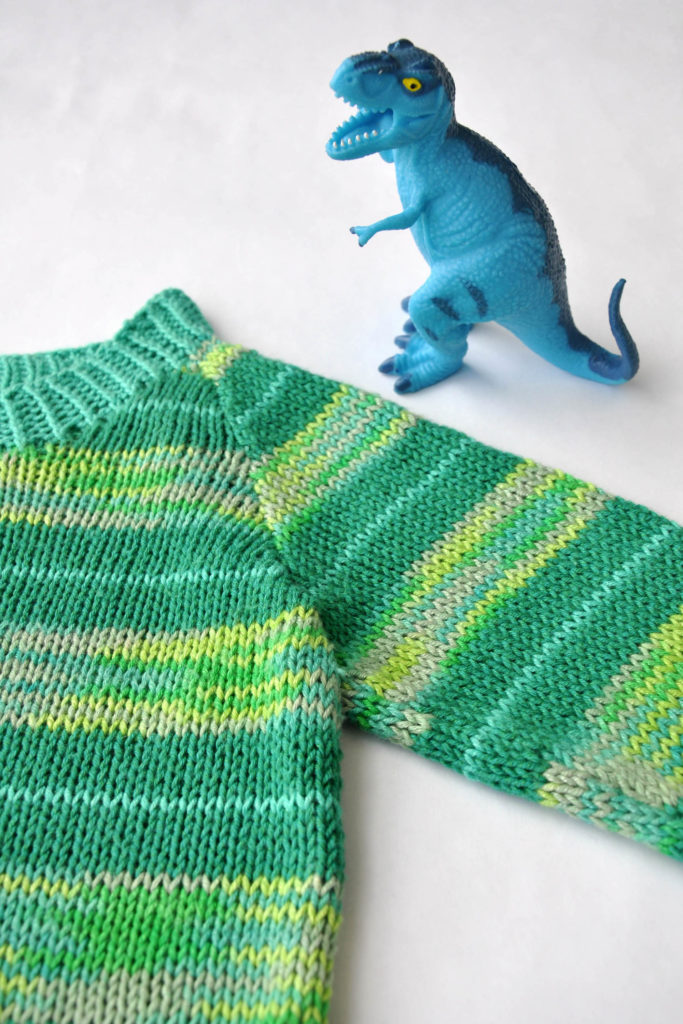 Blue toy dinosaur standing next to shoulder of green children's knit pullover