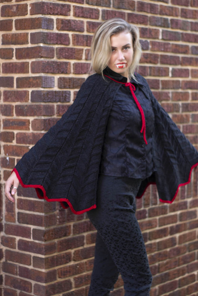 Young woman with vampire fangs wearing a black cape knit in Uptown DK. 
