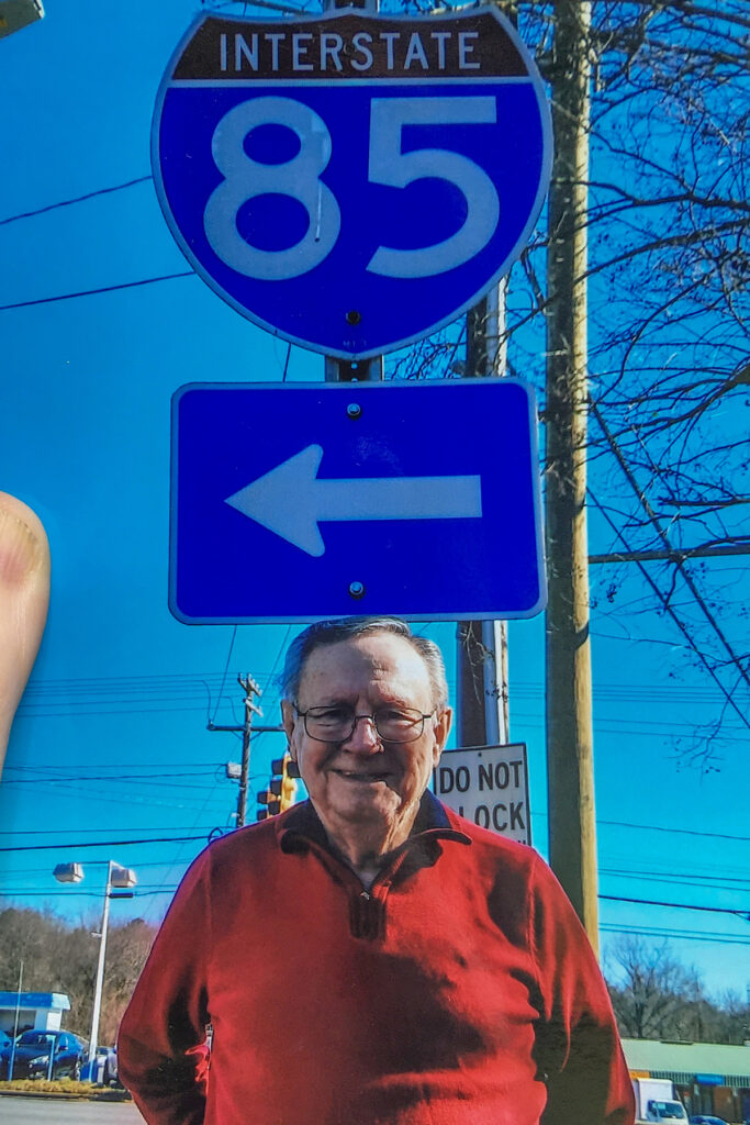 Image of 85-year-old man standing below highway sign reading "Interstate 85"