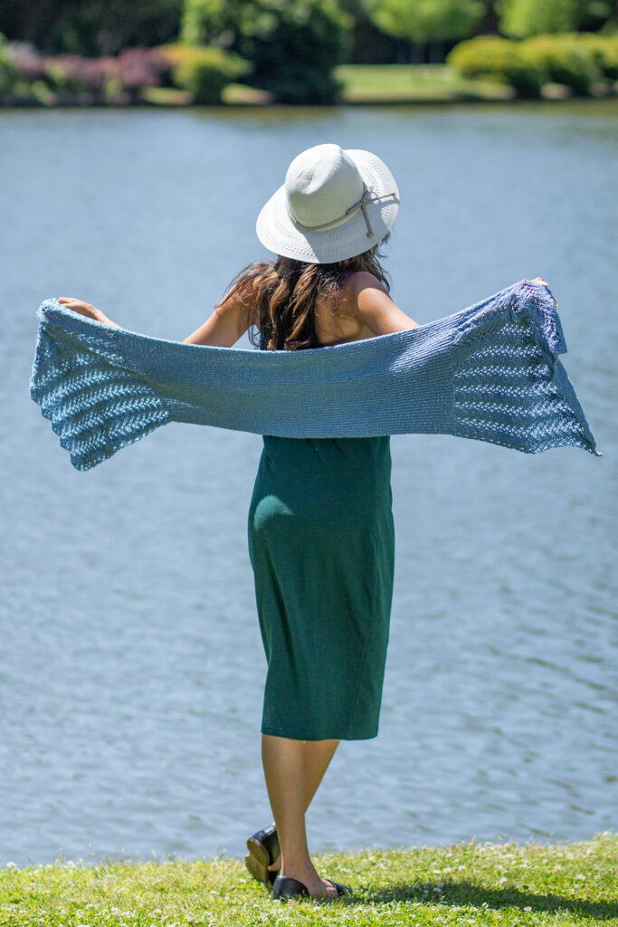 Woman facing away from viewer, holding a light blue knitted wrap outstretched.