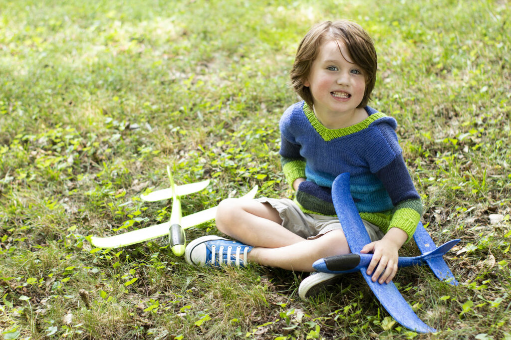 Young boy seated in grass, wearing knitted striped hoodie and holding two toy airplanes