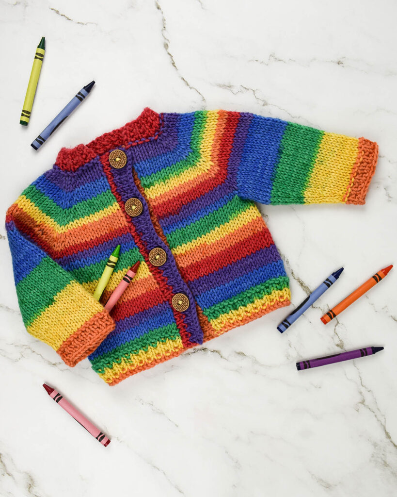 Striped baby cardigan knitted in Deluxe Stripes yarn. Crayons are scattered around it.