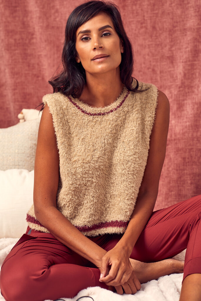 A woman seated on blankets and pillows wearing red pants and a camel-color vest with red details, knit using Universal Yarn Minou. 