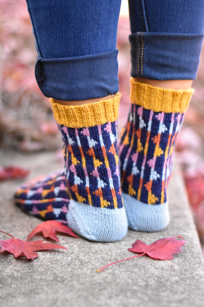 A detailed view of the handknit Party Flag Socks, shown from behind. Maple leaves are lying on the ground next to the person wearing the socks. 