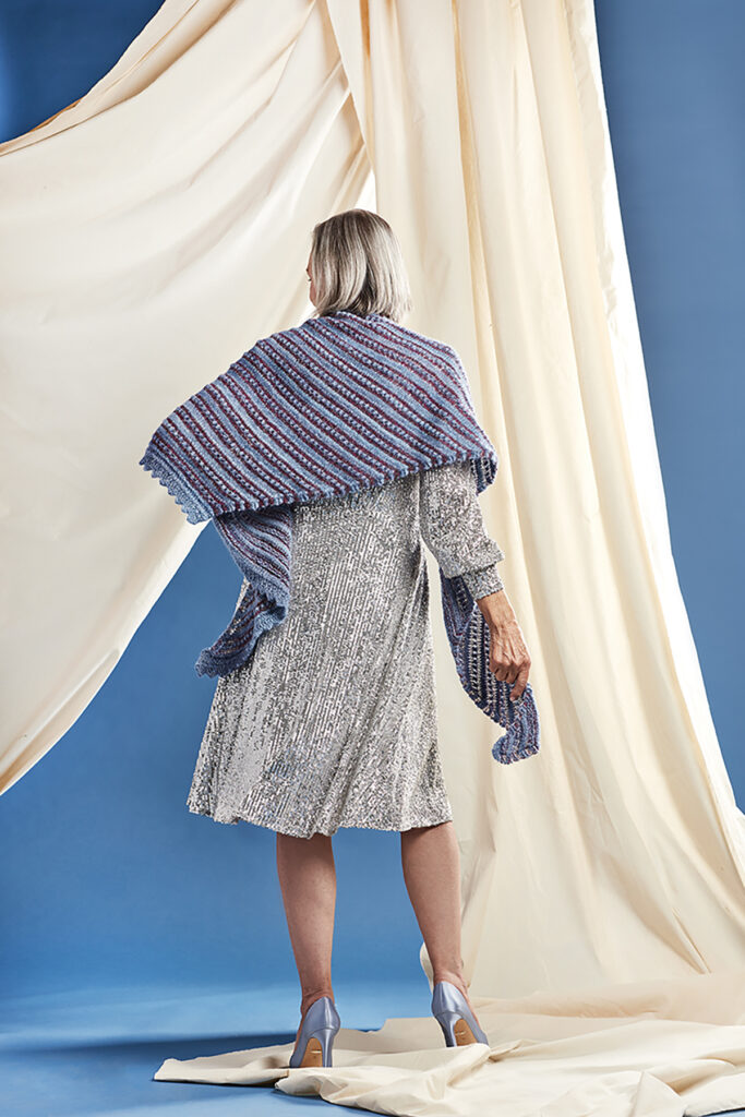 A woman stands while wearing the Ardenne Shawl, hand knitted with Universal Yarn Noelle and Universe. She is shown from the back, and glances down to the left. 
