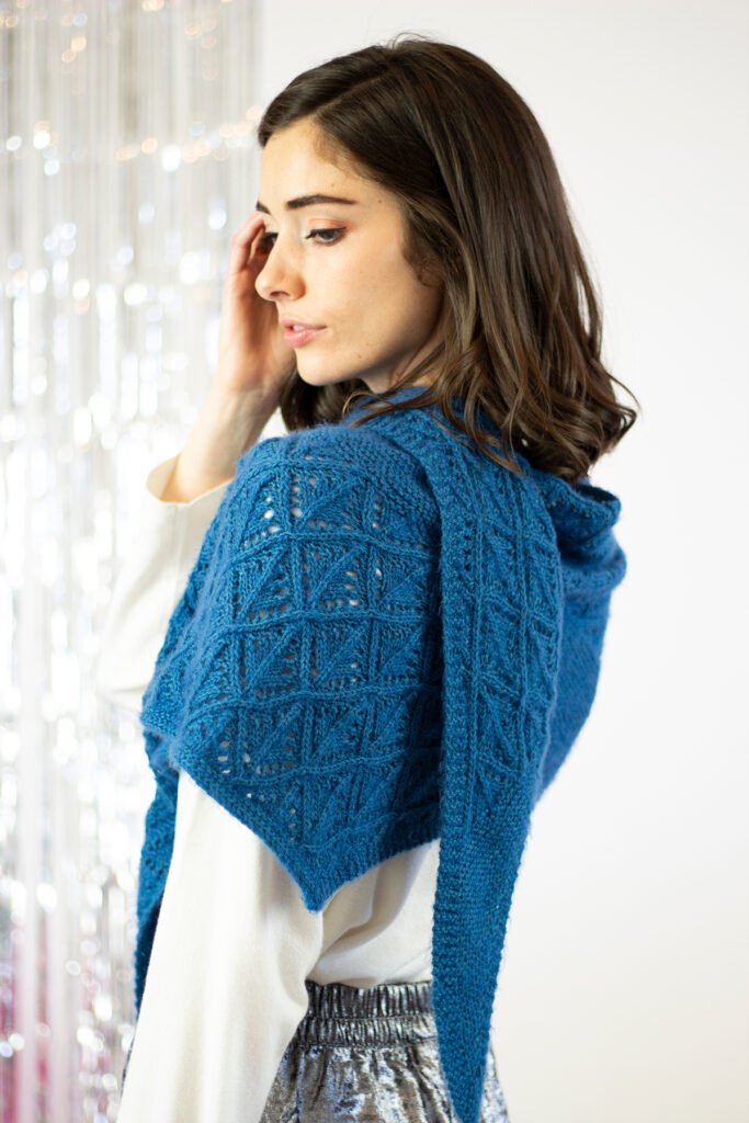 A close up image of a person facing away from the camera and looking down. They are wearing a hand knit shawl in a dark blue color on their shoulders.