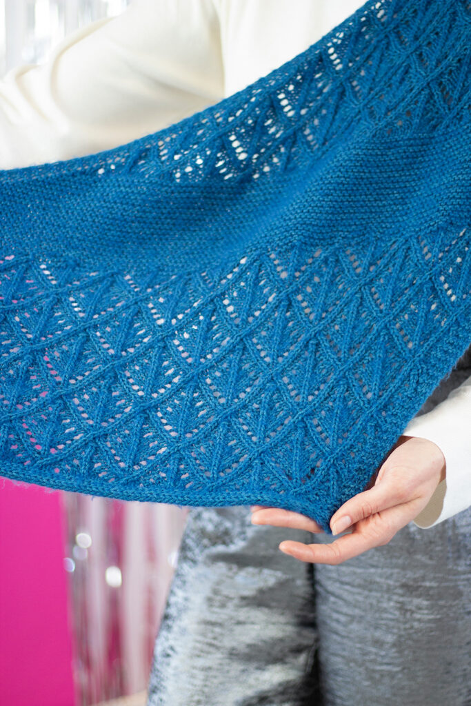 A close up image of the details of a hand knit blue shawl with lace stitches. 
