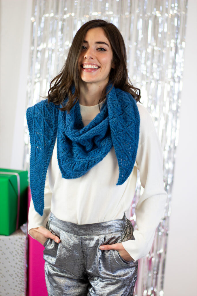 A person wearing a white top, silver pants, and a hand knit blue shawl
