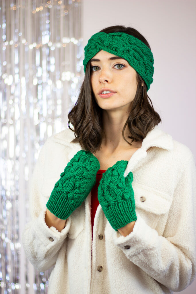 A woman glances directly at the camera while wearing a headband and mittens that are hand knitted using Universal Yarn Deluxe Worsted Superwash. 
