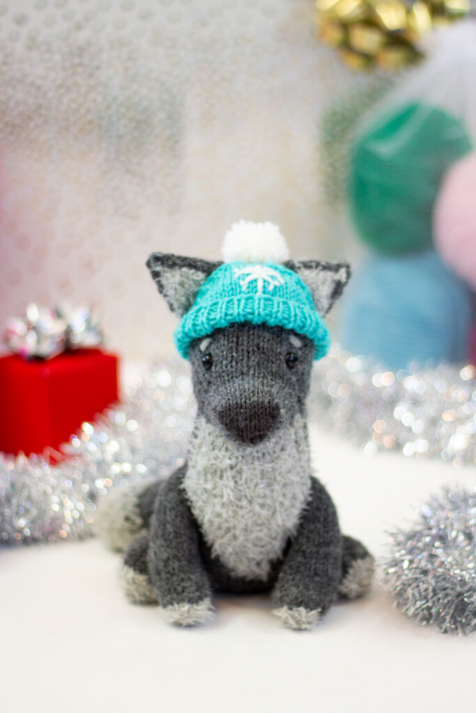 A front view of a small, hand knitted wolf toy using Universal Yarn Minou and Deluxe Worsted. The toy wears an embroidered turquoise hat with a pom pom. 