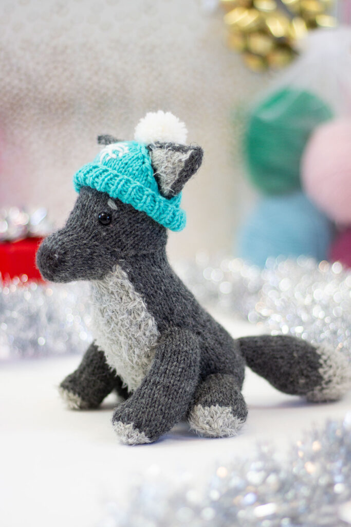 A small, hand knitted wolf toy, shown from the side and wearing a turquoise hat. 