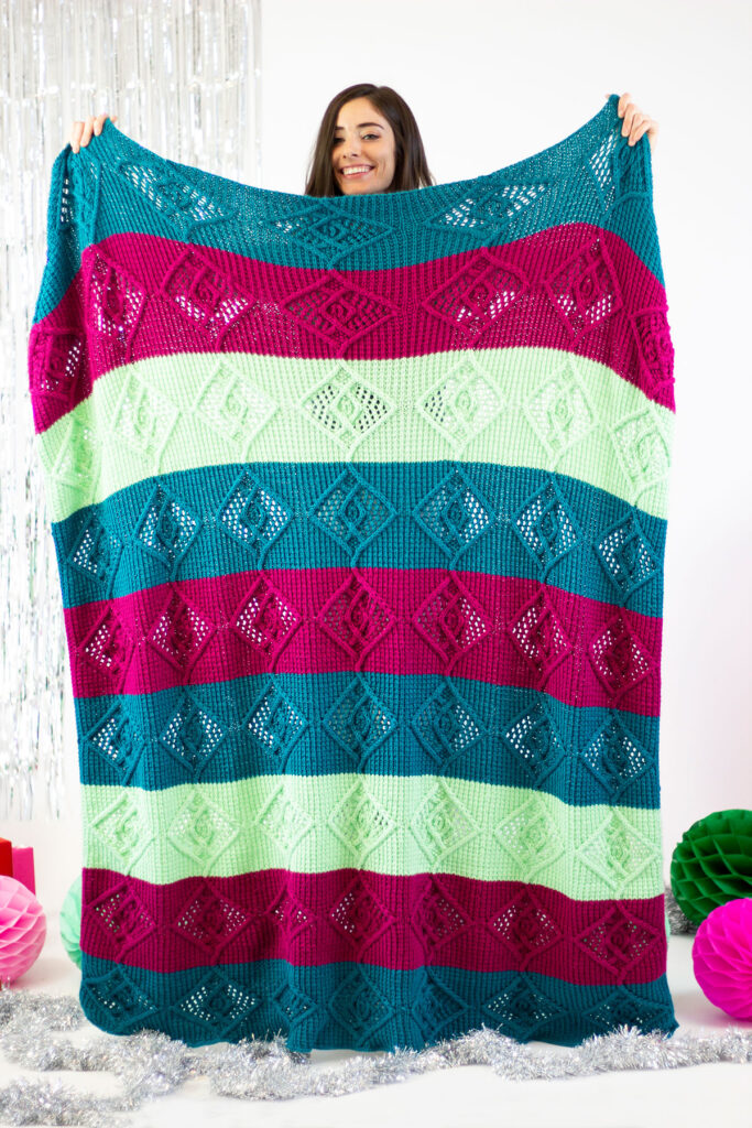 A person stands holding up a large hand knit blanket in a blue, magenta, and green striped colorway. 