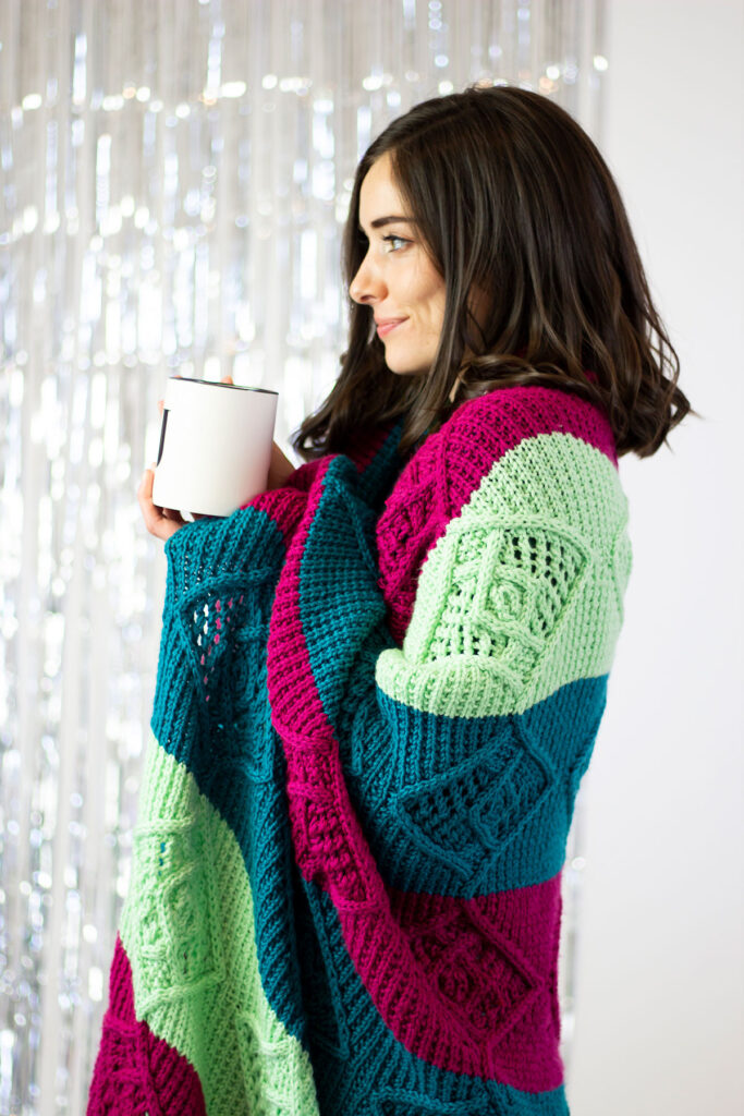 A person standing sideways holding a mug and has a hand knit blanket wrapped around their shoulders.