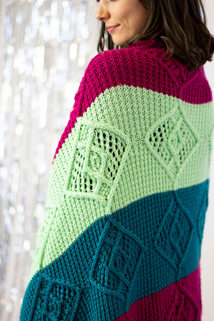 A close up image of the details of a hand knit striped blanket with lace and cable details. 
