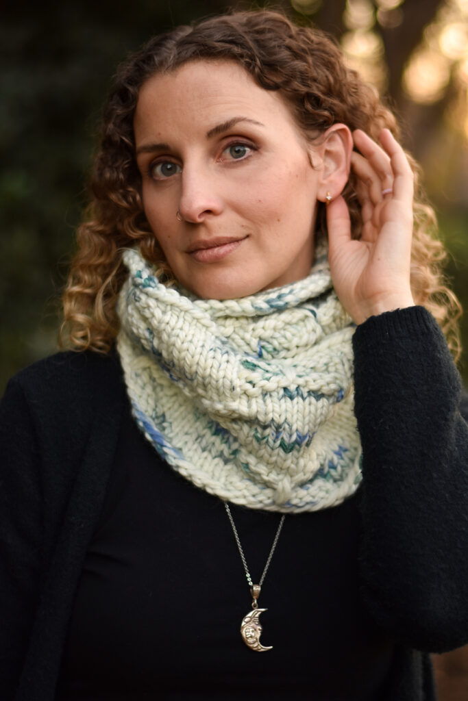 A person wearing a black sweater and a hand knit cowl in a cream, blue and green colorway 
