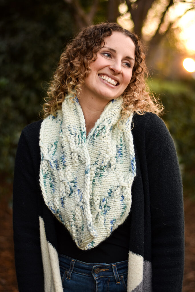 A person standing outside looking away from the camera and smiling. They are wearing a black sweater and a hand knit cowl in a cream and blue colorway. 