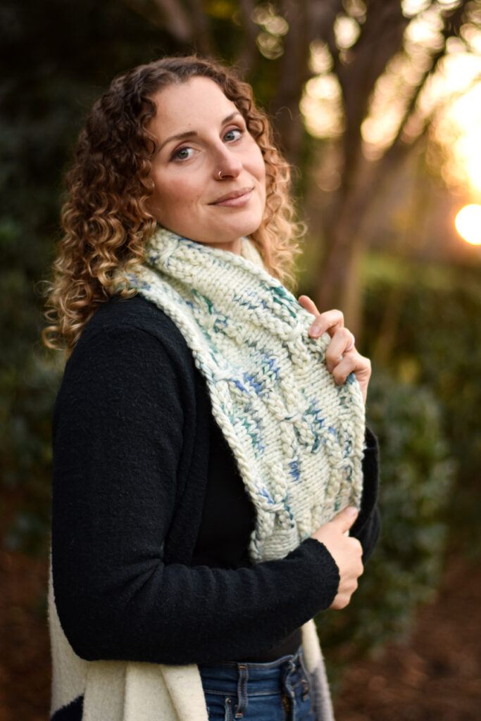 A person standing sideways looking at the camera. They are wearing a black sweater and a hand knit cowl in a cream, blue, and green colorway. 