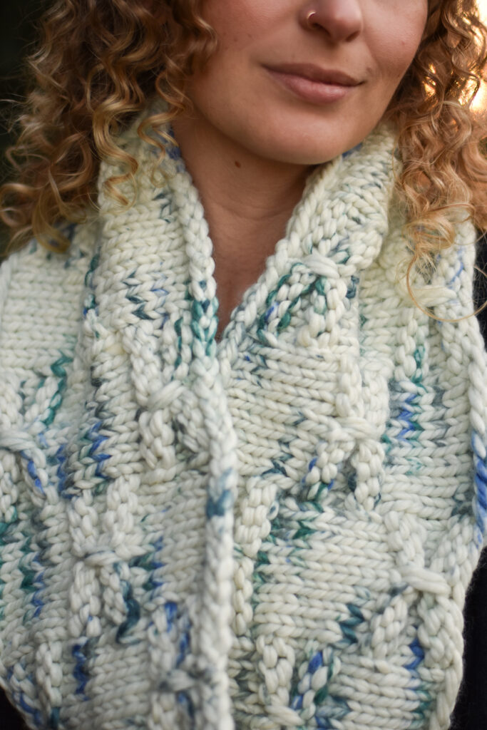 detail shot of a person wearing a hand knit cowl in a cream, blue, and green colorway.