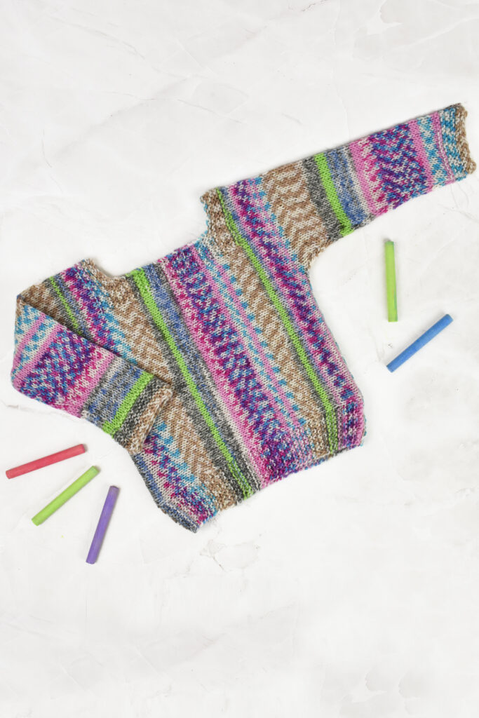 A flat lay of a baby sweater hand knitted with Universal Yarn Easel. It is surrounded with chalk in colors that coordinate with the sweater. 