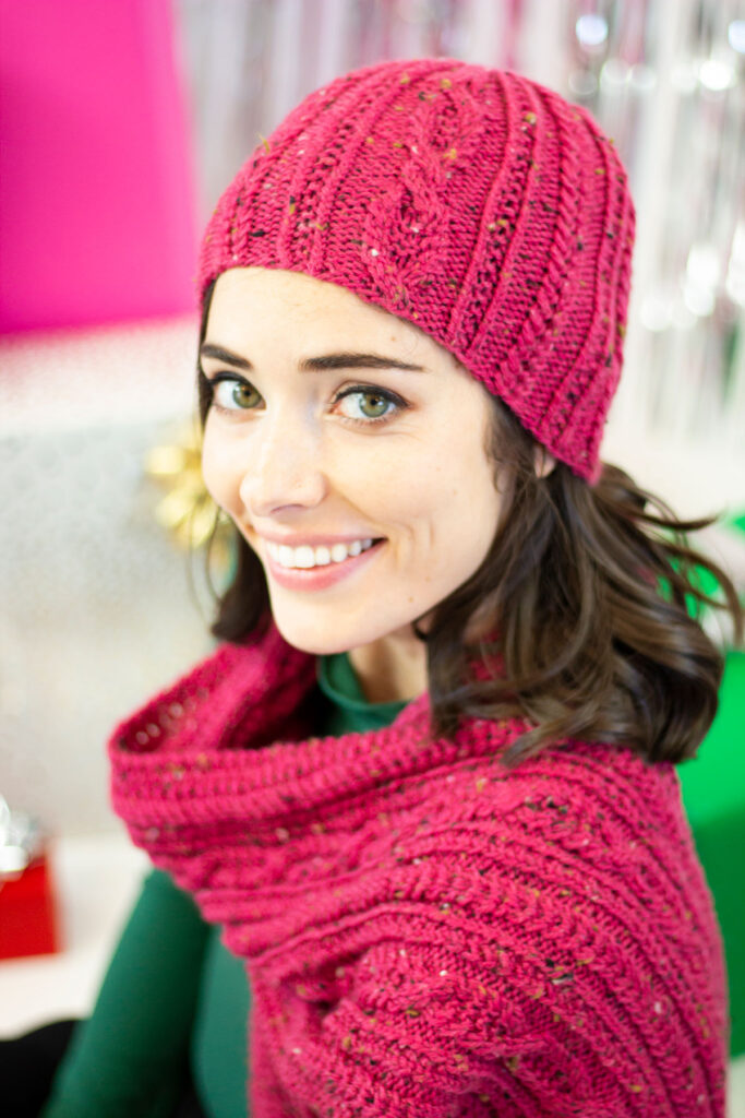 A brunette woman smiles while wearing a hand knit hat and scarf that features cables, twisted stitches, and texture throughout. 
