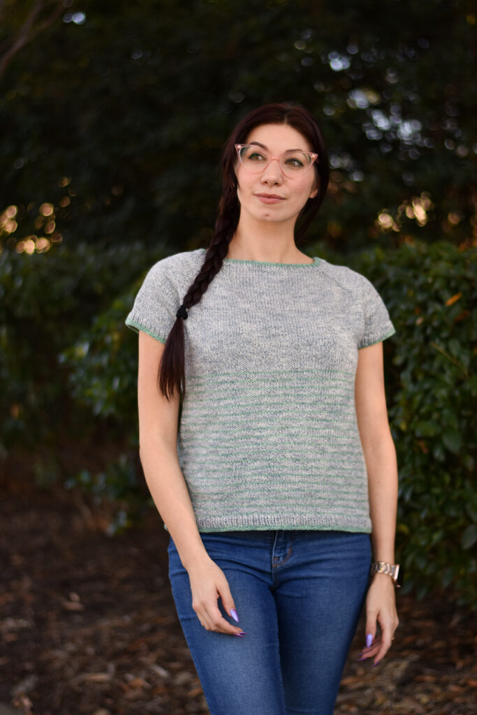 A woman wearing pink glasses stands in front of greenery and is wearing a striped top, handknitted using Universal Yarn Truva. 