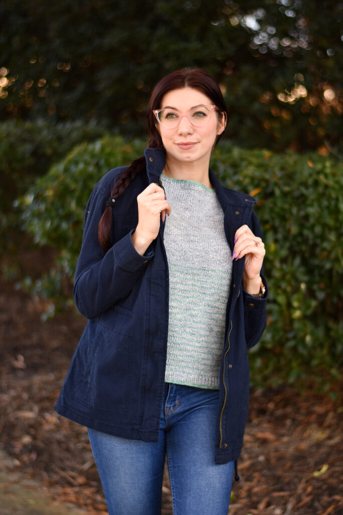 A woman with pink glasses stands in front of greenery wearing a top handknitted using Universal Yarn Truva. She is also wearing a navy blue jacket. 
