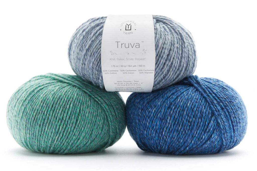 Three balls of Universal Yarn Truva. On the top, a yarn ball has a label and is light blue. It sits on top of a green and darker blue ball of the same yarn. 