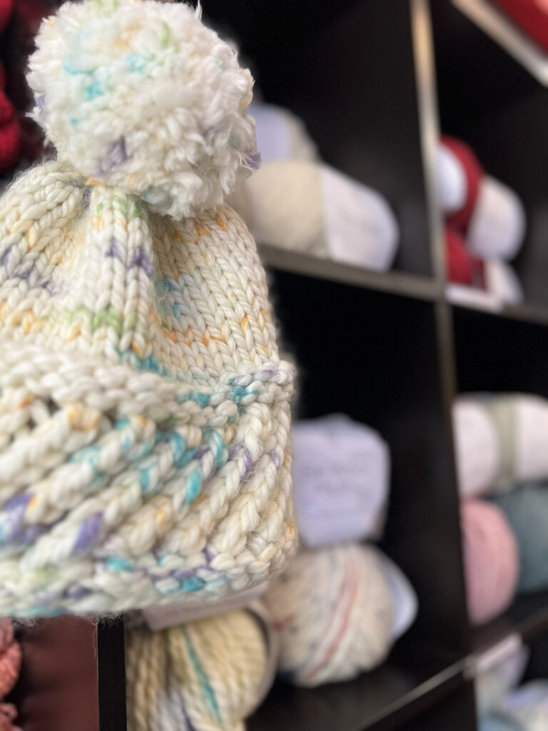 Photo of a hat knit in Be Wool Multis with a shelf of Be Wool Multis and Be Wool in the background