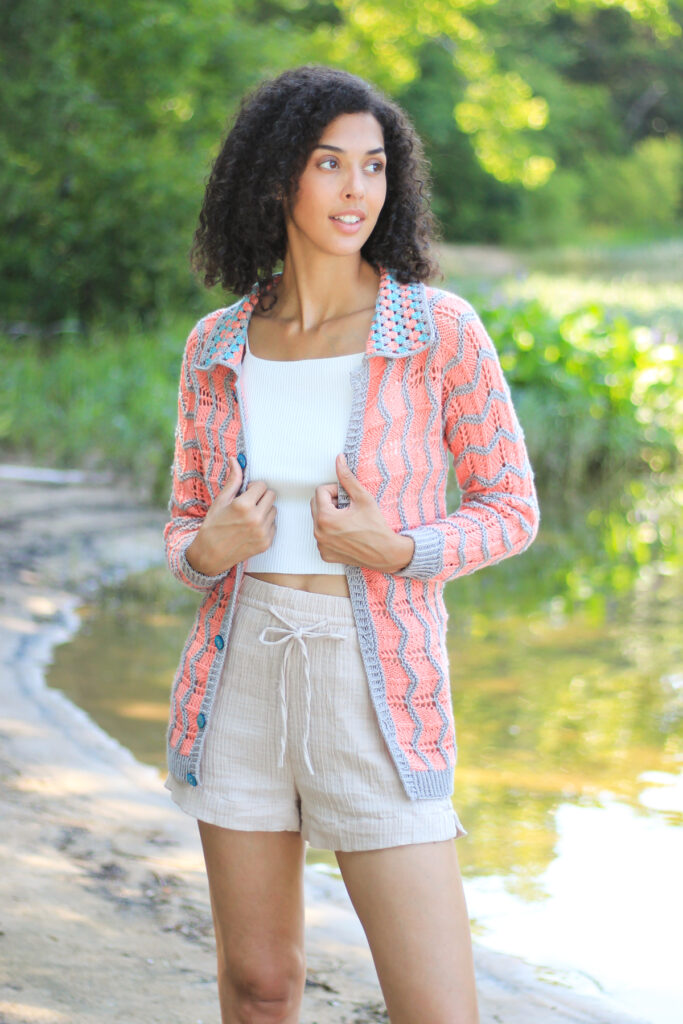 A young woman stands by a lake while wearing linen shorts, a tank, and a hand knit and crochet cardigan made with Universal Yarn Uptown Bamboo DK. 