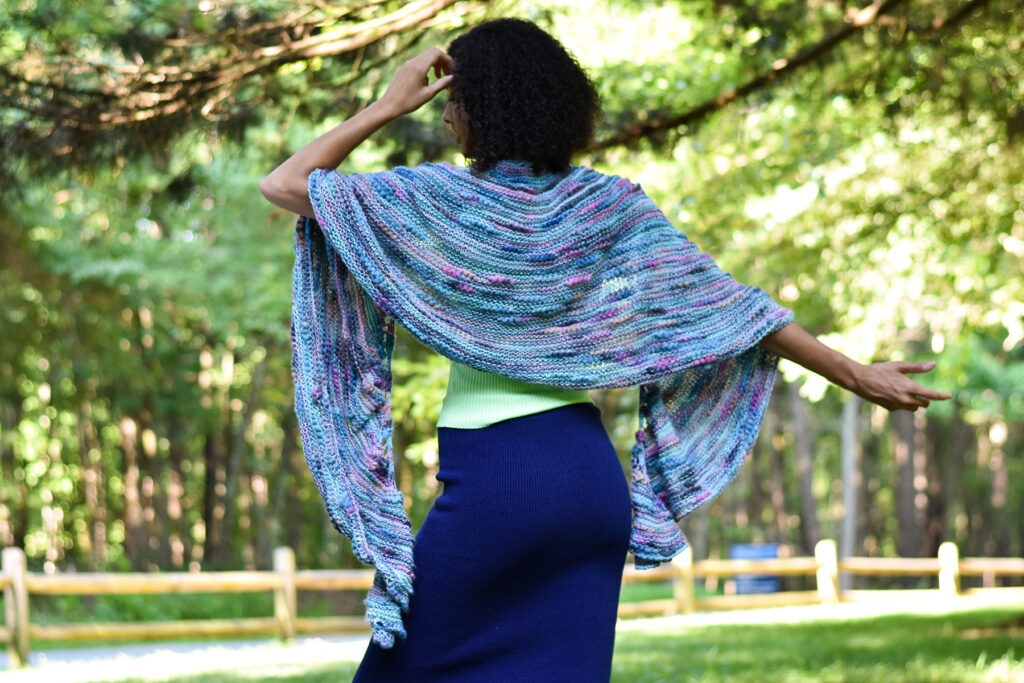 Photo of a person wearing the Kerid shawl facing away from the camera with trees in the background
