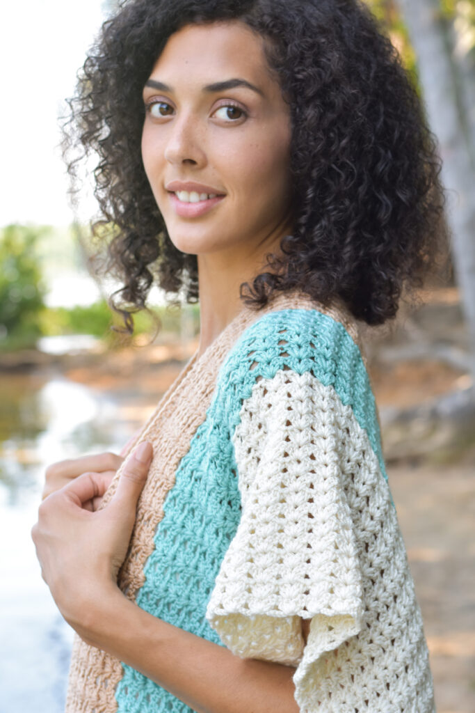 A young woman glances over her shoulder to the camera. She is wearing a crocheted Subtle Shell Ruana, made with Universal Yarn Uptown Bamboo DK. 