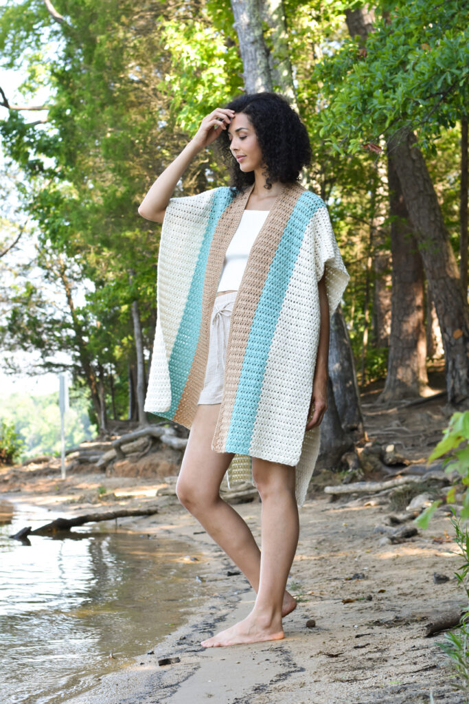 A young woman touches her hair and looks slightly downward, standing by a lake. She is wearing linen shorts, a white tank, and the crocheted Subtle Shell Ruana in Universal Yarn Uptown Bamboo DK. 