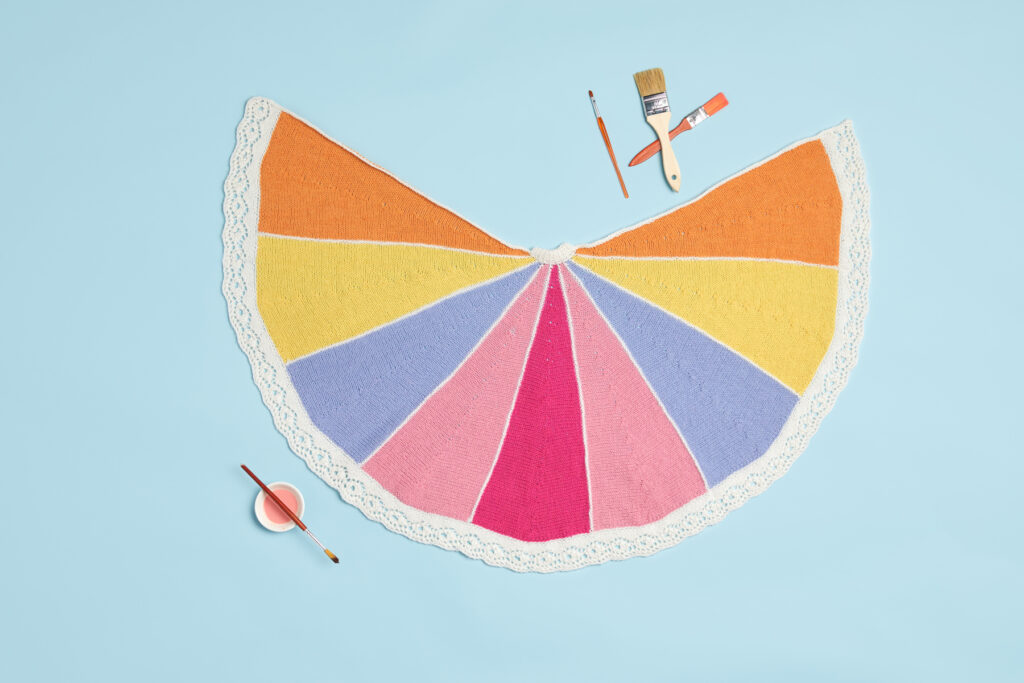 Photo of the Color Wheel Shawl on a blue background with paint brushes and paint